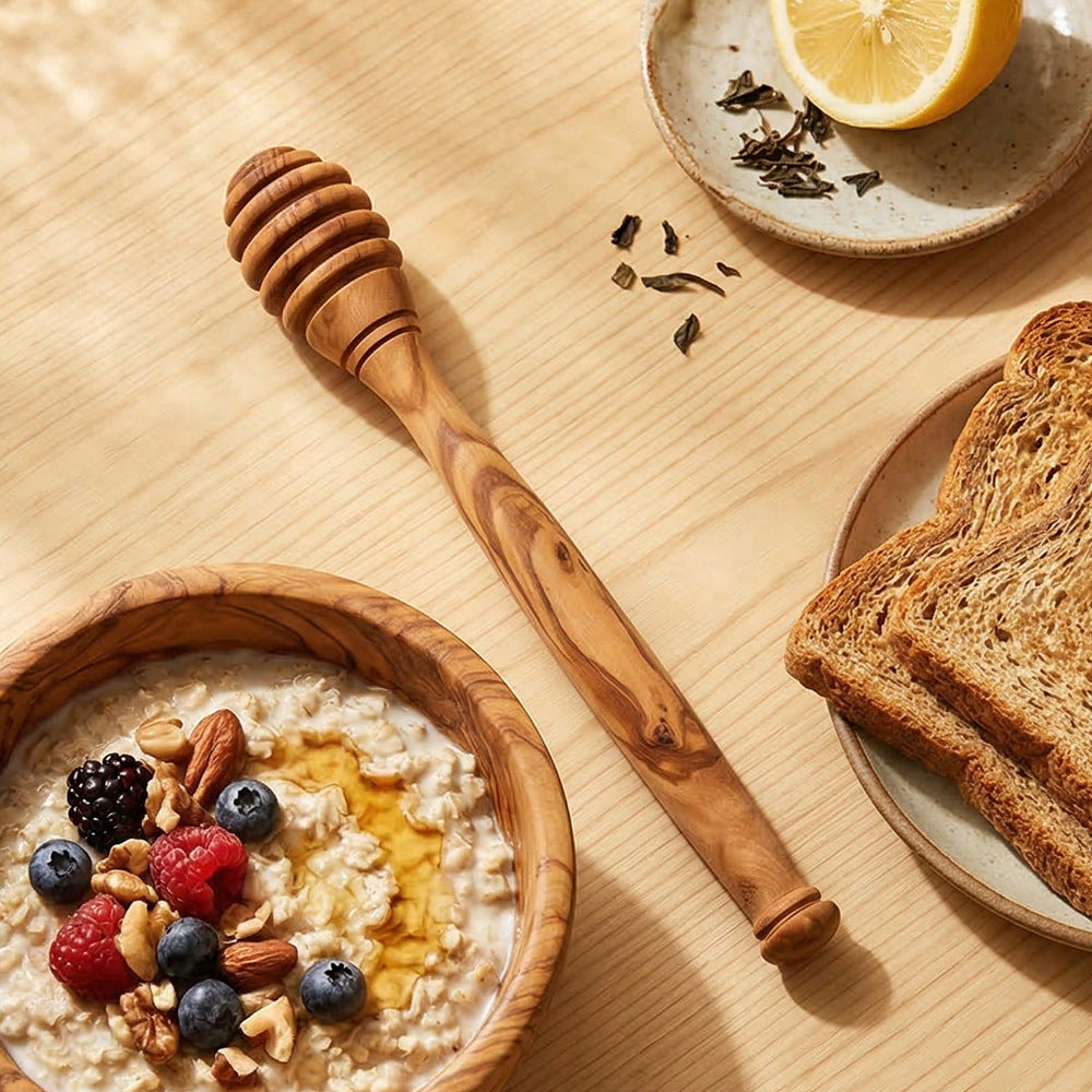 Handcrafted olive wood honey dipper stick with rich natural grain beside a breakfast table.